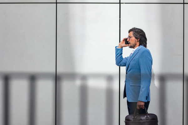 Portrait of mature businessman phone calling, standing on city street, in front of office building. Handsome man with glasses in a suit.