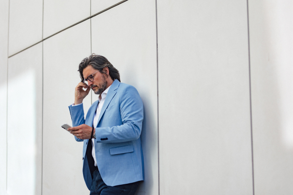 Portrait of mature businessman looking at phone, standing on city street, in front of office building. Handsome man with glasses in a suit.