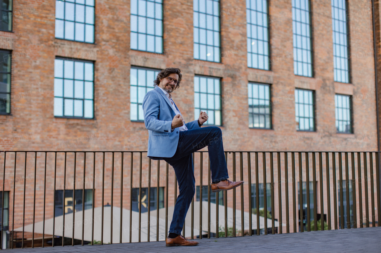 Portrait of happy mature businessman standing on city street, in front of office building. Handsome man with glasses in a suit celebrating successful business deal.