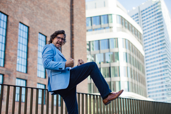 Portrait of happy mature businessman standing on city street, in front of office building. Handsome man with glasses in a suit celebrating successful business deal.