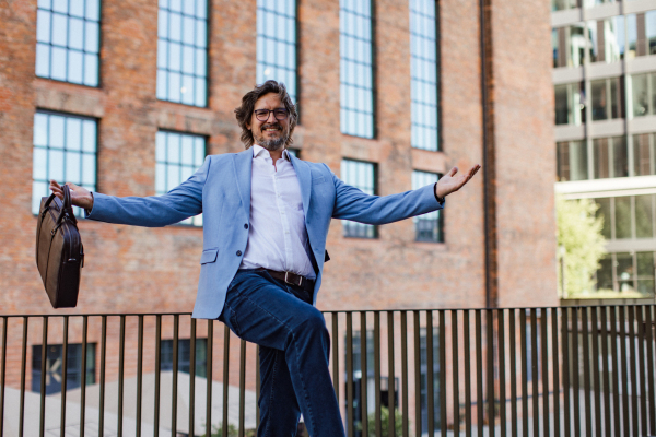 Portrait of happy mature businessman standing on city street, in front of office building. Handsome man with glasses in a suit celebrating successful business deal.