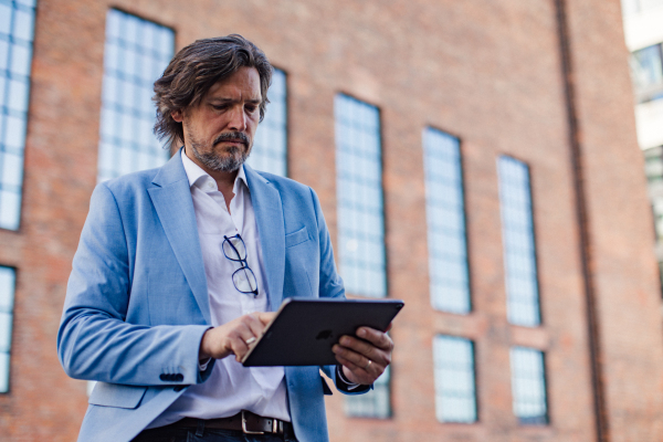 Portrait of happy mature businessman standing on city street, looking at tablet. Handsome man with glasses in a suit celebrating successful business deal.