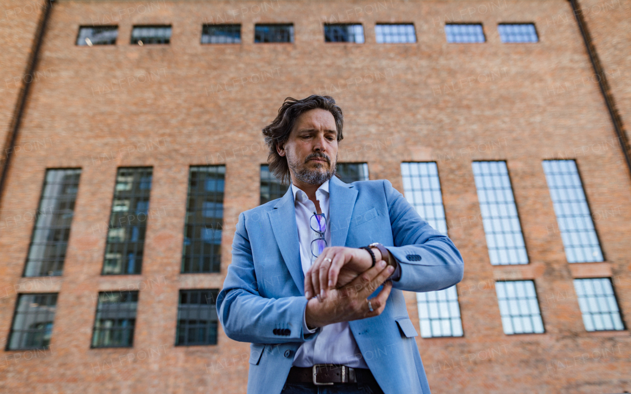 Portrait of mature businessman standing on city street, checking time on watch. Handsome man with glasses in a suit.