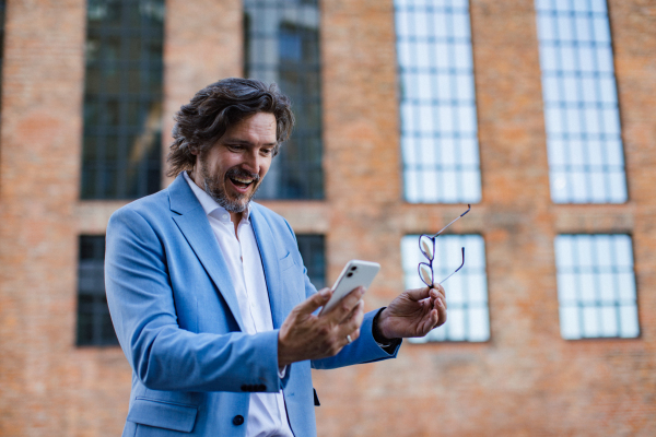 Portrait of happy mature businessman standing on city street, looking at smartphone. Handsome man with glasses in a suit celebrating successful business deal.