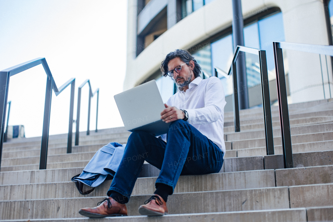 Portrait of mature businessman sitting on stairs in city, working on laptop. Handsome man with glasses in a suit.