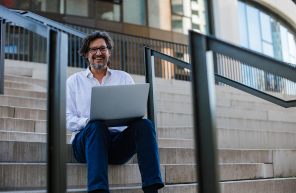 Portrait of mature businessman sitting on stairs in city, working on laptop. Handsome man with glasses in a suit.