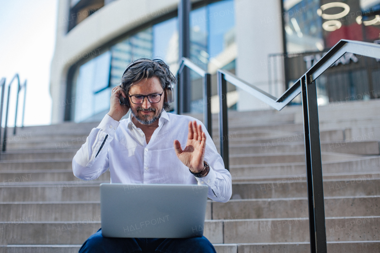 Portrait of mature businessman sitting on stairs in city, working on laptop. Handsome man with glasses in a suit.