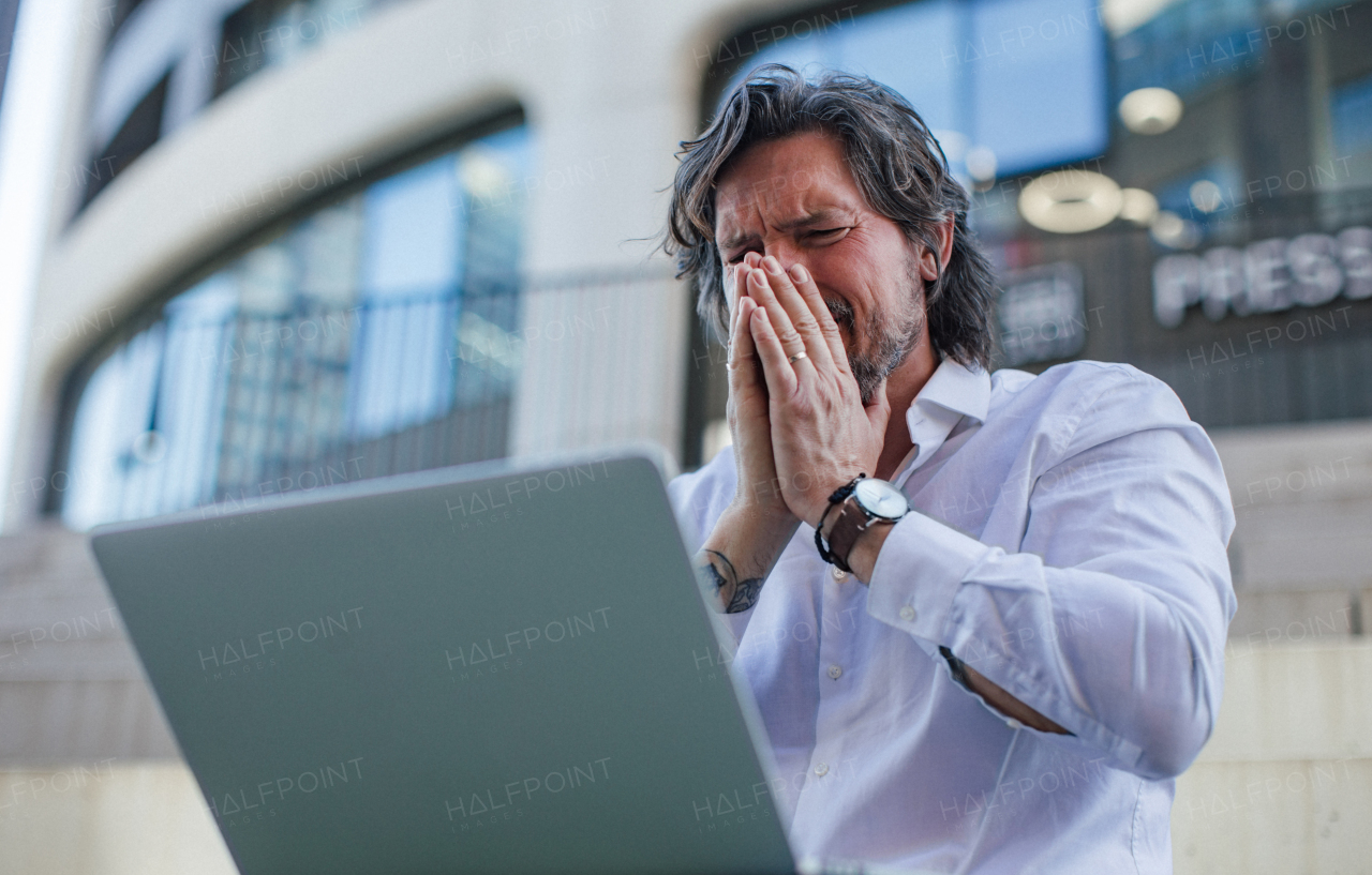 Portrait of mature businessman working on laptop outdoor, recieving bad news. Handsome man with glasses in a suit.