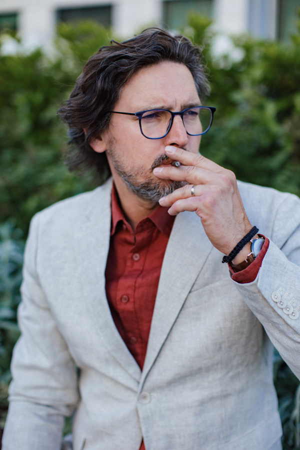 Portrait of mature businessman standing on city street, smoking cigarette. Handsome man with glasses in suit, in front of a office building