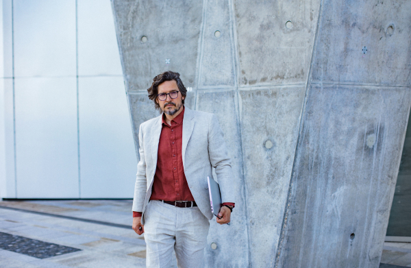 Portrait of mature businessman standing on city street, in front of office building. Handsome man with glasses in a suit.