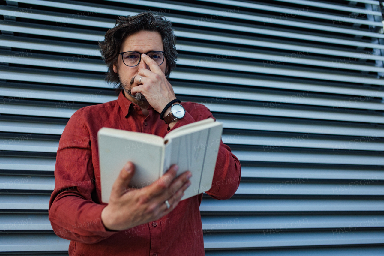 Portrait of mature businessman standing on city street, reading book. Handsome man with glasses in a suit.