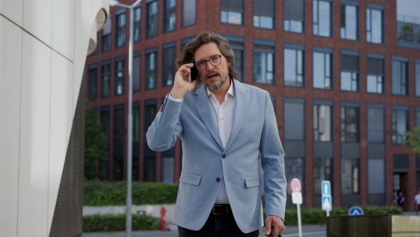 Video of mature businessman phone calling, standing on city street, in front of office building. Handsome man with glasses in a suit.