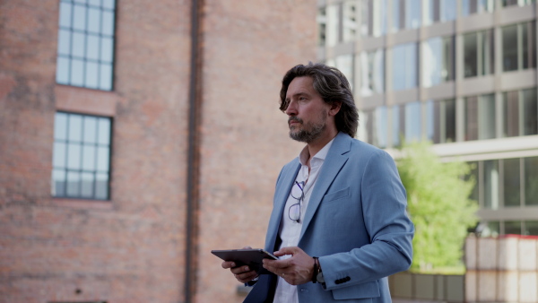 Video of mature businessman with tablet, walking on city street, in front of office building. Handsome man with glasses in sky blue suit.