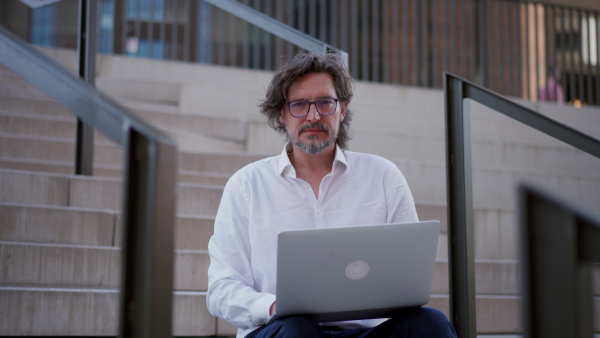 Video of mature businessman sitting on stairs in city, working on laptop. Handsome man with glasses in a suit.