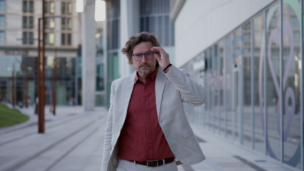 Video of mature businessman phone calling, walking on city street, in front of office building. Handsome man with glasses in a suit.