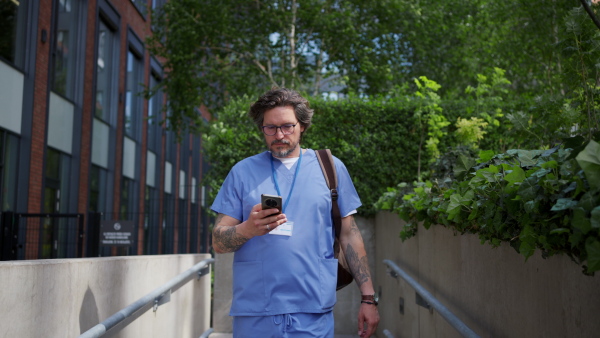 Video of handsome doctor in blue uniform outdoors, scrolling phone. Work-life balance of healthcare worker.