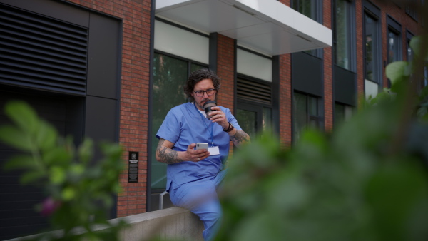 Healthcare worker taking a break, scrolling a phone and drinking coffee outdoors, in front of the hospital building.