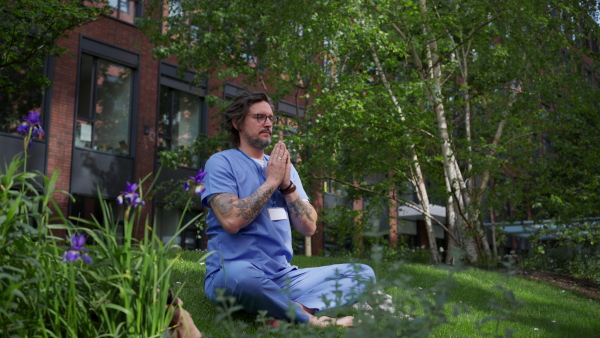 Handsome doctor sitting on grass, meditating and relaxing, winding down from busy workday in hospital. Mental health and life balance for a healthcare worker.