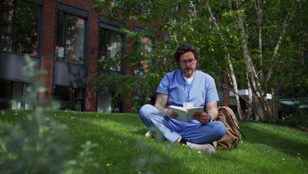 Handsome doctor sitting on grass, reading book and relaxing, winding down from busy workday in hospital. Mental health and life balance for a healthcare worker.