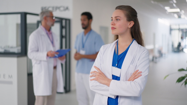 Young woman doctor at hospital corridor, looking at camera, her colleagues standing and talking in the background.