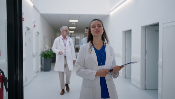 Young woman doctor walking on hospital corridor, with the colleagues in the background.