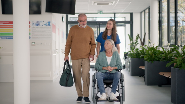 Young nurse pushing wheelchair and talking with senior patients at hospital corridor.