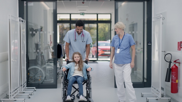 Young caregiver pushing little girl on wheelchair at a hospital corridor, having fun together.