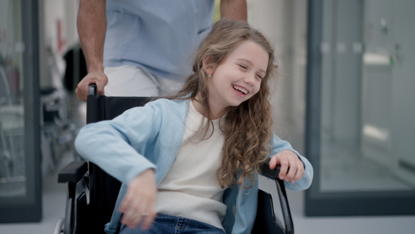 Young caregiver pushing little girl on wheelchair at a hospital corridor, having fun together.