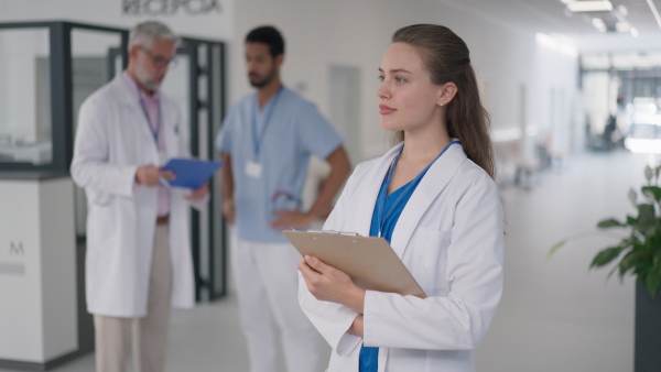 Young woman doctor at hospital corridor, looking at camera, her colleagues standing and talking in the background.