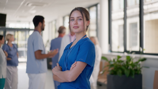 Young woman doctor at hospital corridor, looking at camera, her colleagues standing and talking in the background.