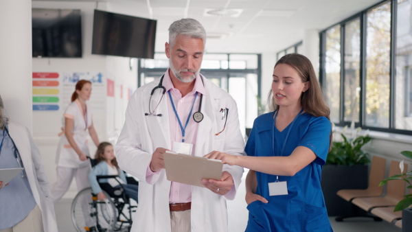 Older doctor giving advise to younger colleague, discussing at hospital corridor. Health care concept.