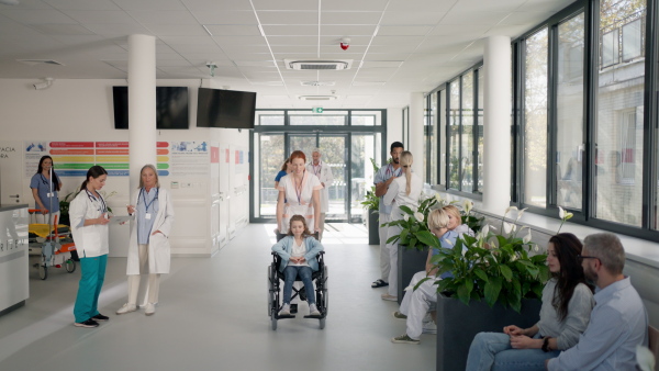 Young nurse pushing little girl on wheelchair at a hospital corridor.