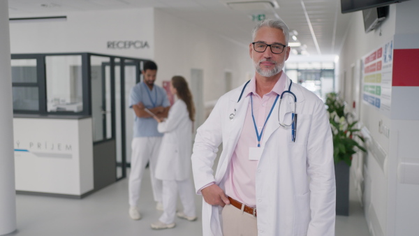 Close-up of mature doctor standing on hospital corridor, looking at a camera.