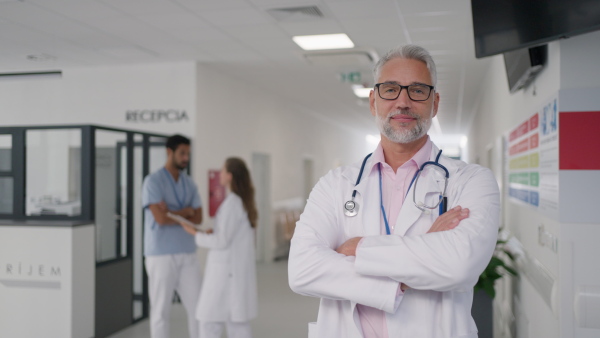 Mature doctor at hospital corridor, looking at camera, her colleagues standing and talking in the background.