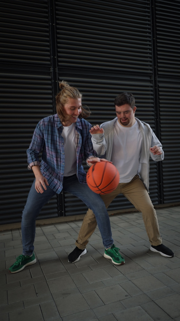 Vertical video of man with down syndrome playing basketball outdoor with his friend. Concept of friendship and integration people with disability into the society.