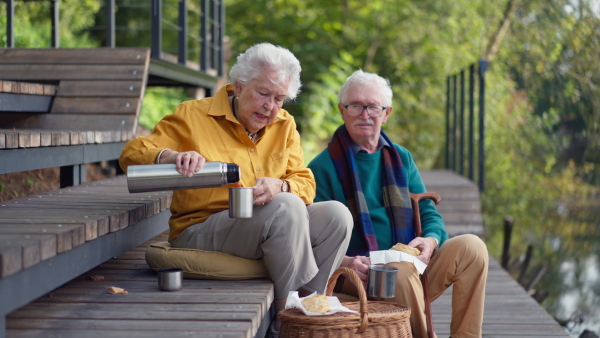 Happy senior couple in autumn clothes having break near a lake after walk.