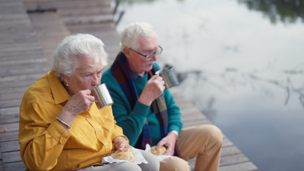 Happy senior couple in autumn clothes having break near a lake after walk.