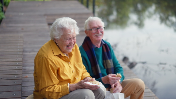 Happy senior couple in autumn clothes having break near a lake after walk.