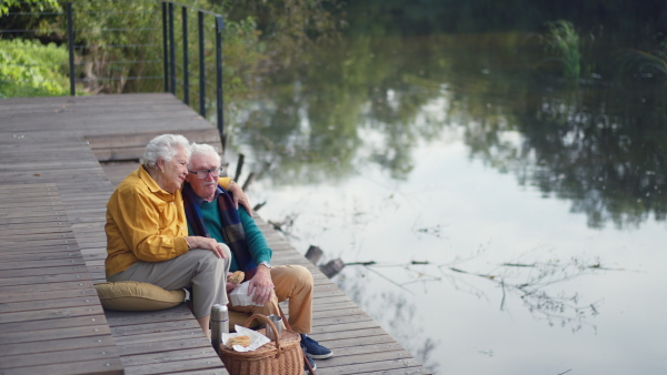 Happy senior couple in autumn clothes having break near a lake after walk.