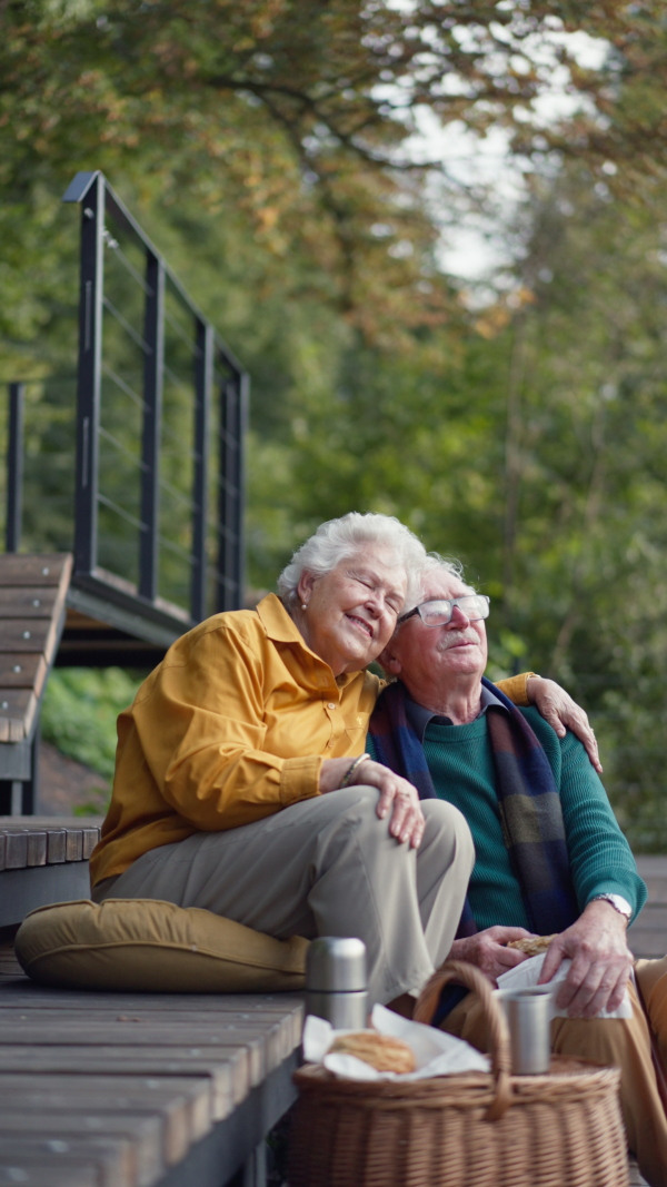 Happy senior couple in autumn clothes having a break and hugging near lake after walk. Vertical footage.