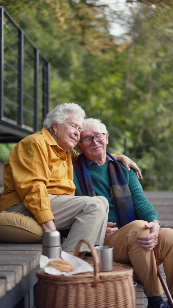 Happy senior couple in autumn clothes having a break and hugging near lake after walk. Vertical footage.