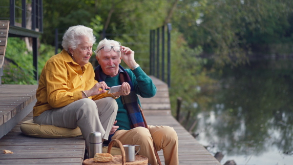 Happy senior couple resting near lake after walk and looking at a smartphone.