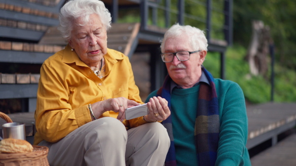 Happy senior couple resting near lake after walk and looking at a smartphone.