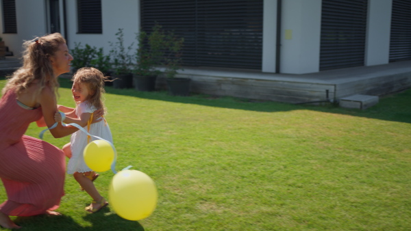 Cute little girl running with yellow balloons in garden, her mother raising her in arms.