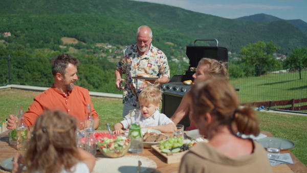 A happy senior man grilling and serving burgers at multi generation garden party in summer.