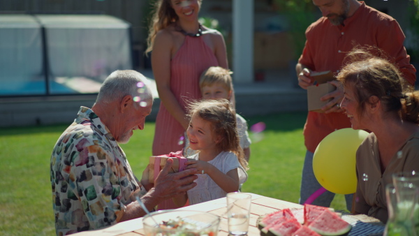 A happy little girl giving birthday present to her senior grandfather at generation family birthday party in summer garden