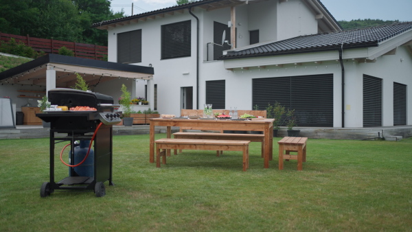 Grill with prepared meat and table with lots of food in garden, in front of house, prepared for garden party, barbecue. Nobody.