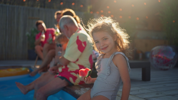 A multi generation family enjoy watermelon and drinks near backyard pool.