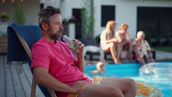 Mature man enjoying summer drink next to swimming pool with family in the background.