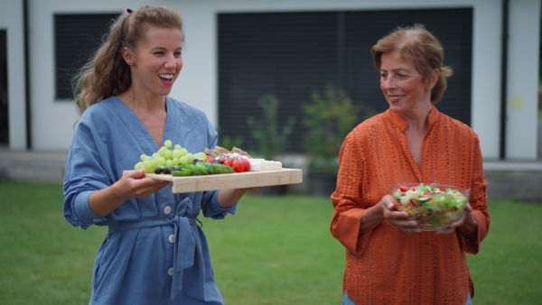 A happy senior woman with her daughter serving salad at multi generation garden party in summer.